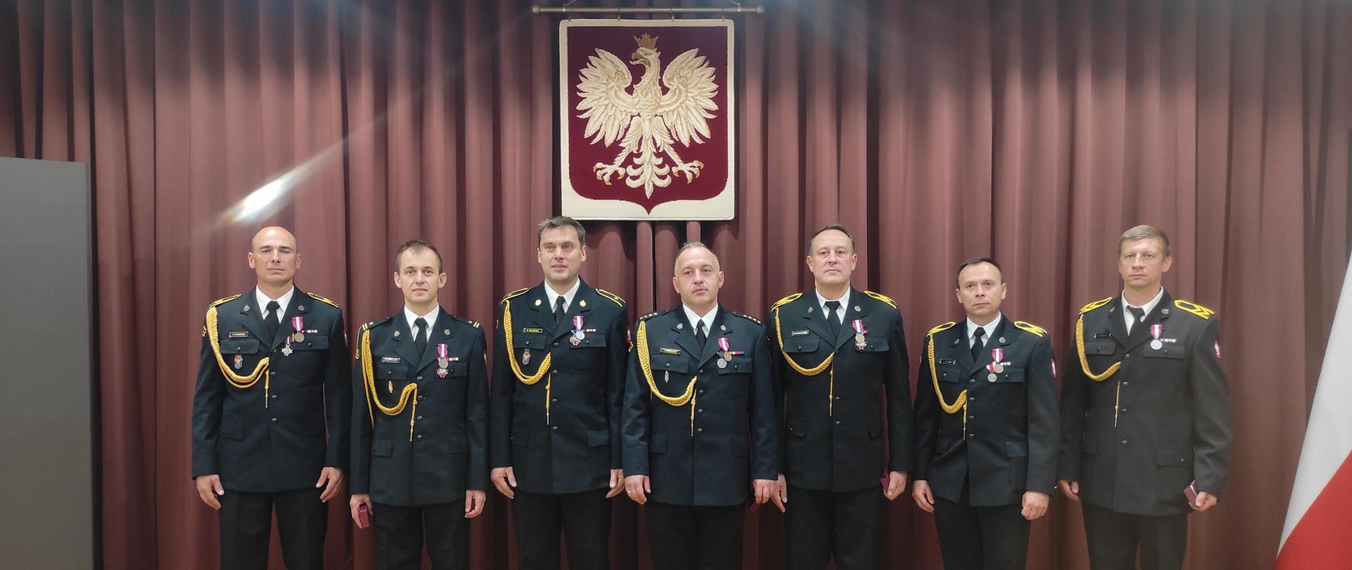 Zdjęcie przedstawia odznaczonych strażaków z KP PSP Sokołów Podlaski ubranych w mundury wyjściowe. Stoją w jednym szeregu na tle bordowej kotary. Nad nimi widnieje orzeł w koronie, godło Polski. Po prawej stronie widzimy biało-czerwoną flagę Polski.