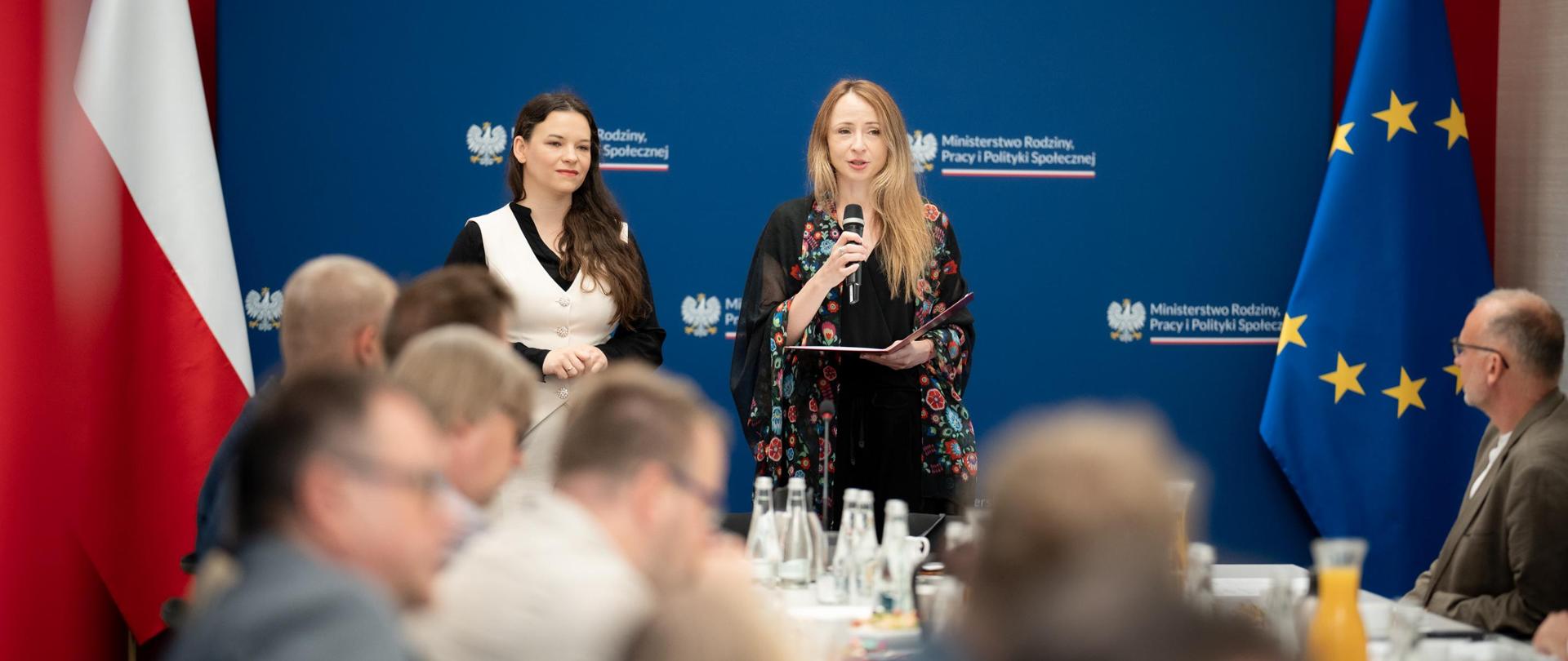 Zdjęcie ministry Dziemianowicz-Bąk oraz wiceministry Nowakowskiej, przemawiających podczas inauguracji zespołu. Obie stoją za stołem przy którym odbywają się obrady, na tle flag i ścianki MRPiPS. W pierwszym planie głowy uczestników zespołu. 