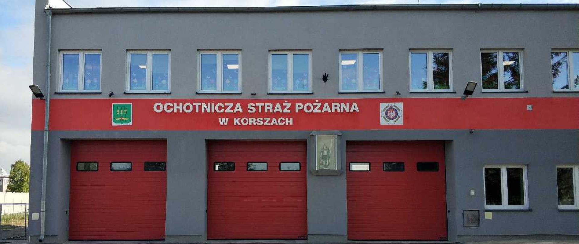 Zdjęcie remizy OSP Korsze, szara elewacja z czerwonym paskiem z napisem Ochotnicza Straż Pożarna w Korszach napisane białą czcionką. 3 czerwone bramy garażowe.