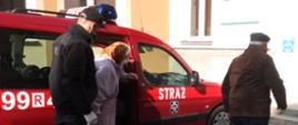 Zdjęcie przedstawia plac na którym zlokalizowany jest punkt szczepień w miejscowości Rakszawa. Strażak Ochotniczej Straży Pożarnej w Rakszawie pomaga małżeństwu w starszym wieku w wyjściu z samochodu pożarniczego. Osoby przemieszczają się do budynku punktu szczepień.
