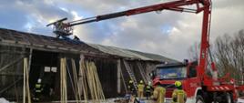 Zdjęcie przedstawia strażaków którzy przy pomocy podnośnika koszowego naprawiają zerwany dach na budynku gospodarczym. 