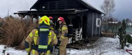 strażacy ubrani w aparaty ochrony układu oddechowego, przygotowują się do wejścia do domku letniskowego. 
