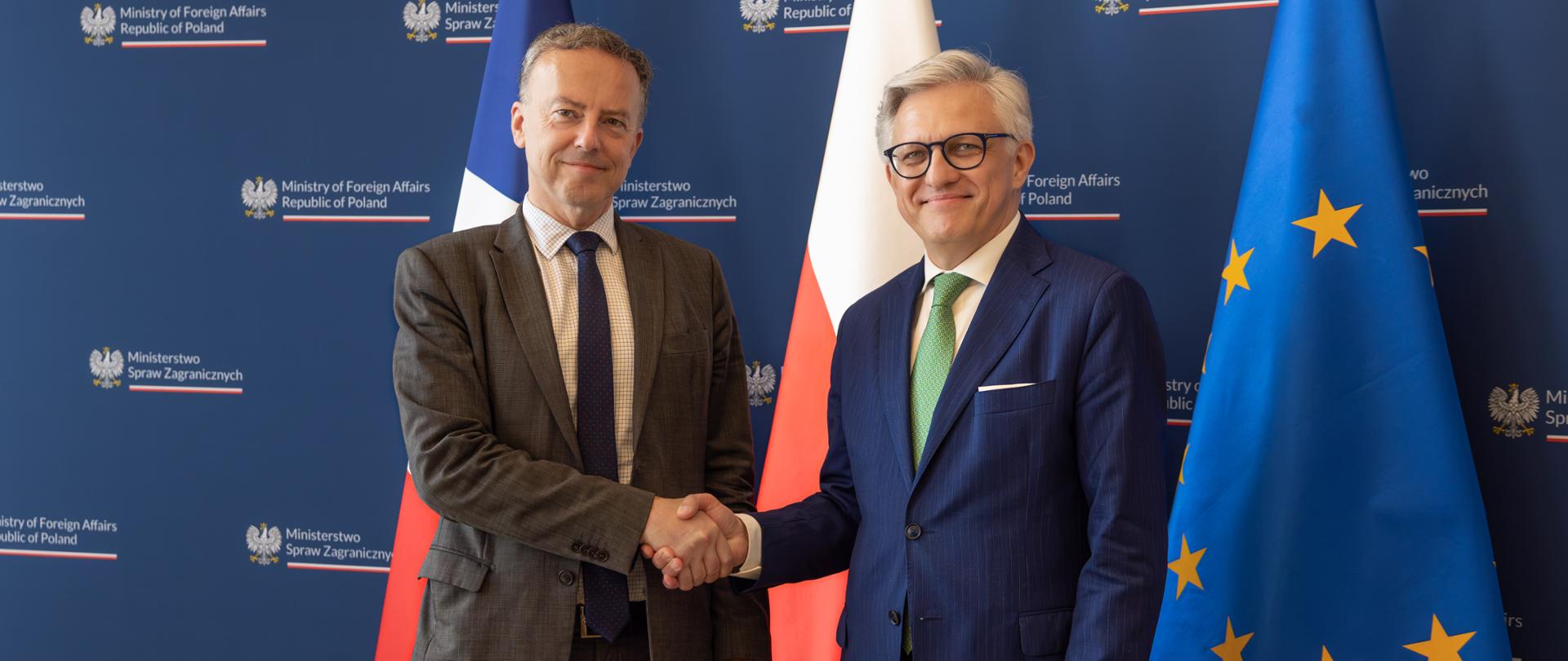 Sekretarz Generalny Ministerstwa Europy i Spraw Zagranicznych Francji Martin Briens i Podsekretarz Stanu Artur Harazim podają sobie ręce na tle granatowej ścianki z logotypami Ministerstwa Spraw Zagranicznych RP oraz flag Francji, Polski i Unii Europejskiej. Obaj uśmiechają się do zdjęcia.