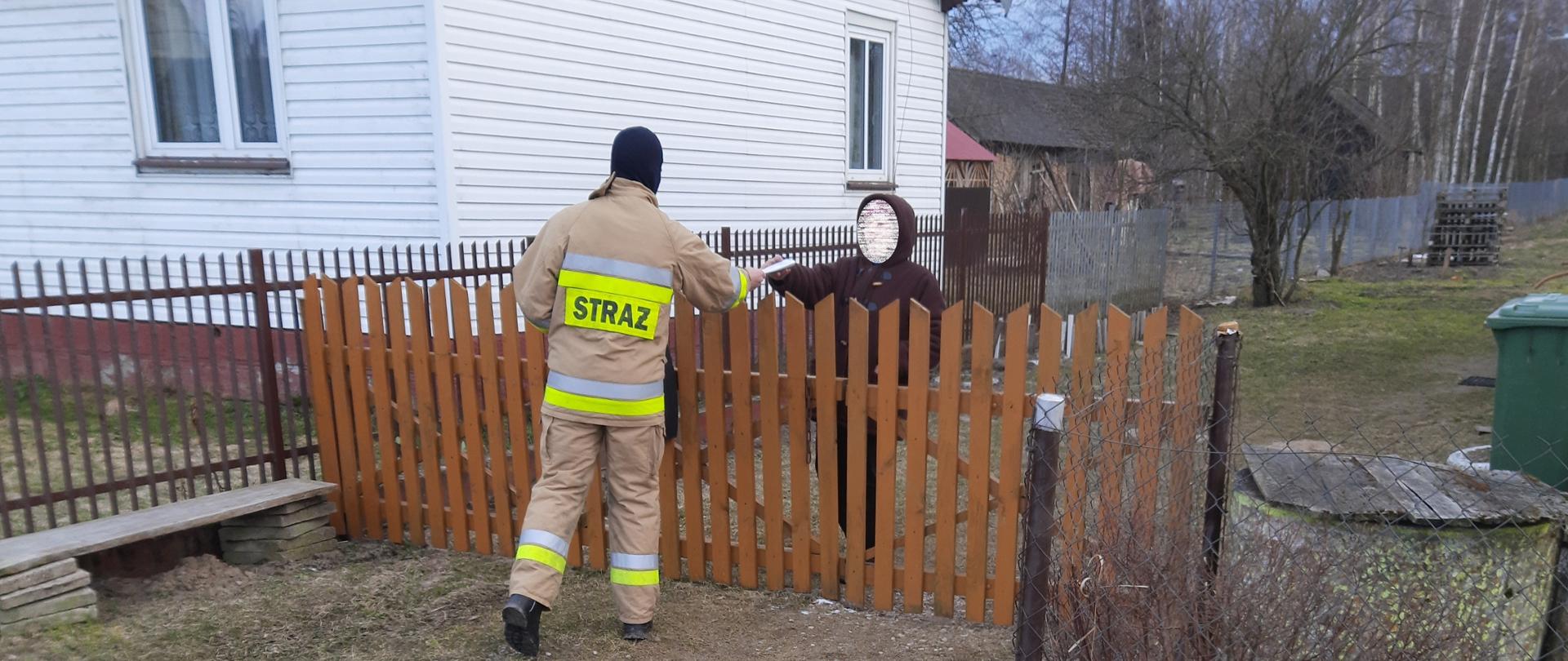Strażak OSP podczas akcji dystrybucji ulotek informacyjnych na temat profilaktyki oraz szczepień przeciwko COVID-19. Strażak przekazuje ulotkę mieszkance jednej z miejscowości powiatu bielskiego.