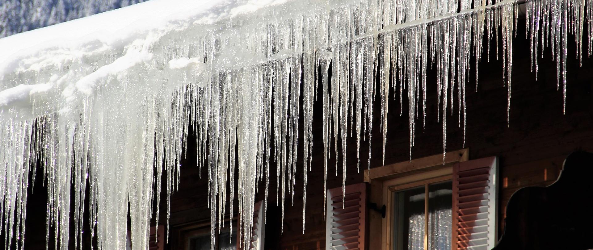 Zdjęcie przedstawia wiszące sople lodu na dachu drewnianego budynku