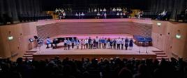 21 dzieci stoi w rzędzie na estradzie sali koncertowej, po lewej stronie kobieta gra na fortepianie, z przodu widać od tyłu publiczność na widowni, nad sceną również widać siedzącą publiczność.