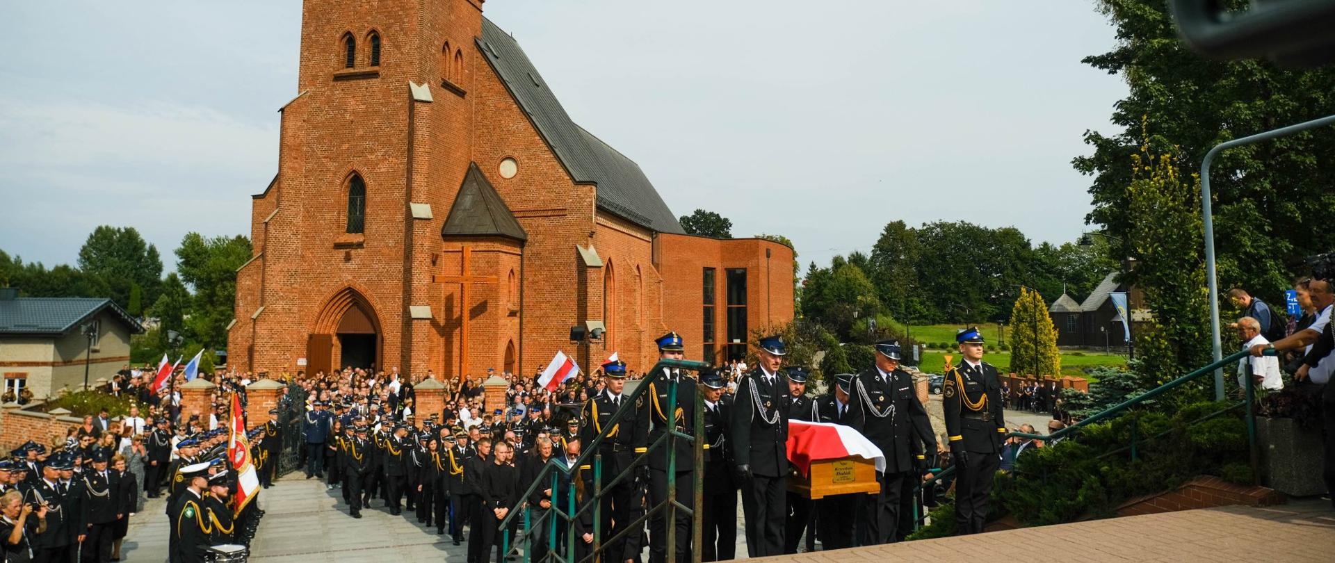 Strażacy niosą trumnę, w tle kompanie reprezentacyjne, poczty sztandarowe straży pożarnej oraz kościół w Sierakowicach