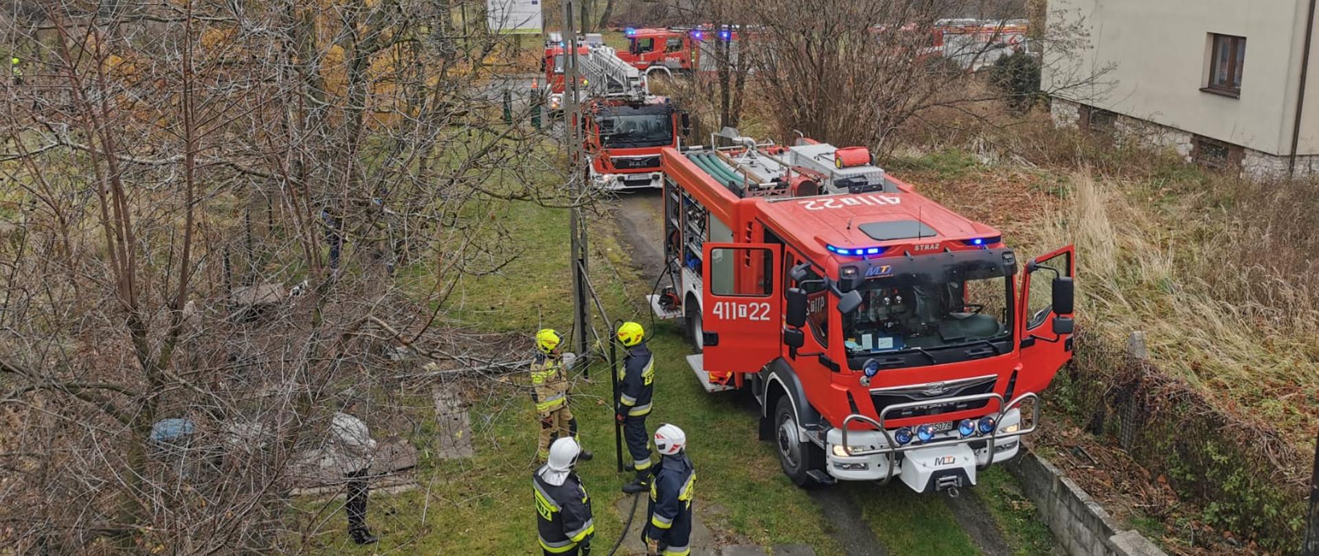 Zdjęcie przedstawia kilka samochodów ratowniczo-gaśniczych ustawionych na drodze wjazdowej do posesji. Przy samochodach znajdują się strażacy w umundurowaniu specjalnym