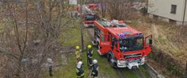 Zdjęcie przedstawia kilka samochodów ratowniczo-gaśniczych ustawionych na drodze wjazdowej do posesji. Przy samochodach znajdują się strażacy w umundurowaniu specjalnym