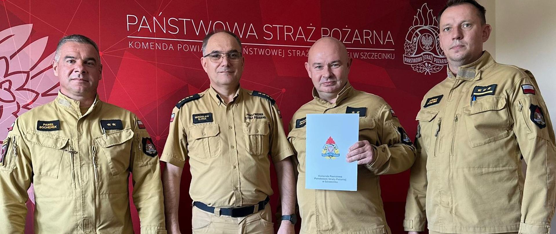 Grupa czterech strażaków w mundurach służbowych stoi razem w pomieszczeniu biurowym, ustawiona w półokręgu. Wszyscy majaą ubrane na sobie jasnobrązowe mundury. W centrum grupy znajduje się strażak trzymający jasną teczkę z dokumentem. W tle logo Państwowej Straży Pożarnej. Wszyscy patrzą w stronę aparatu i uśmiechają się lub zachowują neutralny wyraz twarzy, scena rozgrywa się podczas wręczenia nagrody jubileuszowej.