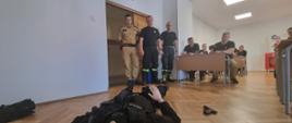 Na podłodze leży manekin ubrany w mundur policyjny przed nim stoi trzech strażaków Państwowej Straży Pożarnej za nimi siedzą w ławkach strażacy biorący udział w szkoleniu. 