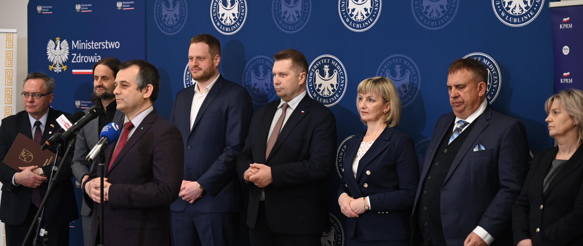 Pod białą ścianą przy której stoją niebieskie banery stoi grupa elegancko ubranych ludzi, jeden mężczyzna mówi do mikrofonu na stojaku.