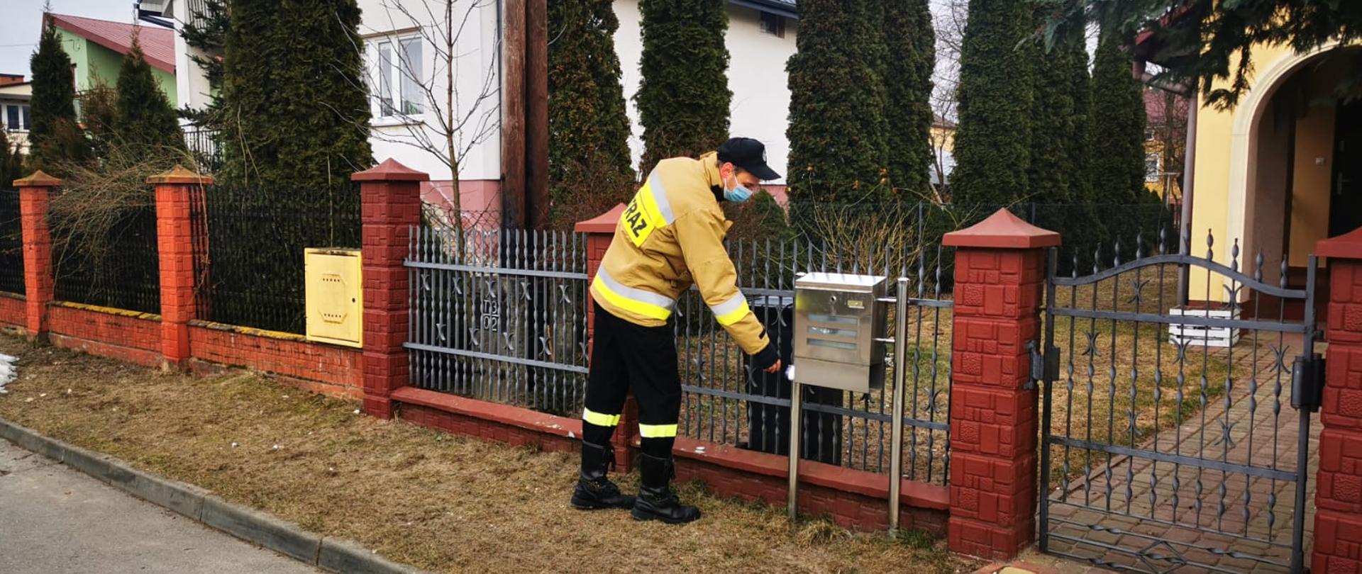 Zdjęcie przedstawia druha Ochotniczej Straży Pożarnej, który wkłada ulotkę do skrzynki pocztowej. W tle dom jednorodzinny.