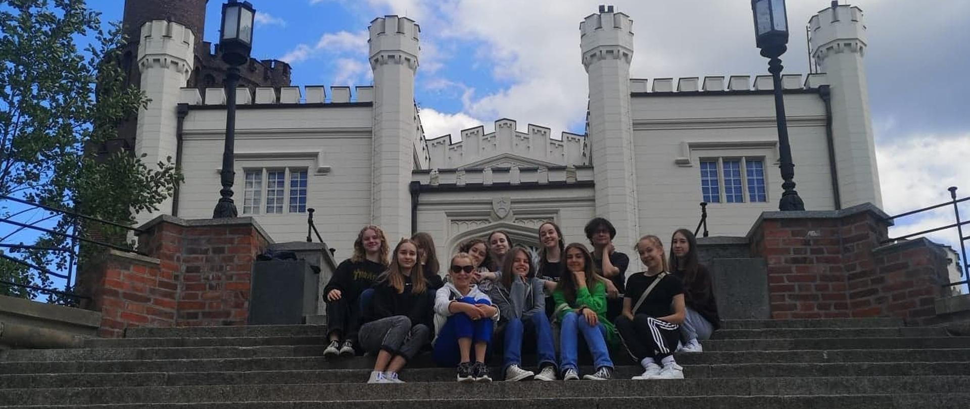 Uczniowie siedzący na schodach budowli w poznaniu (biała przypominająca zamek ) 