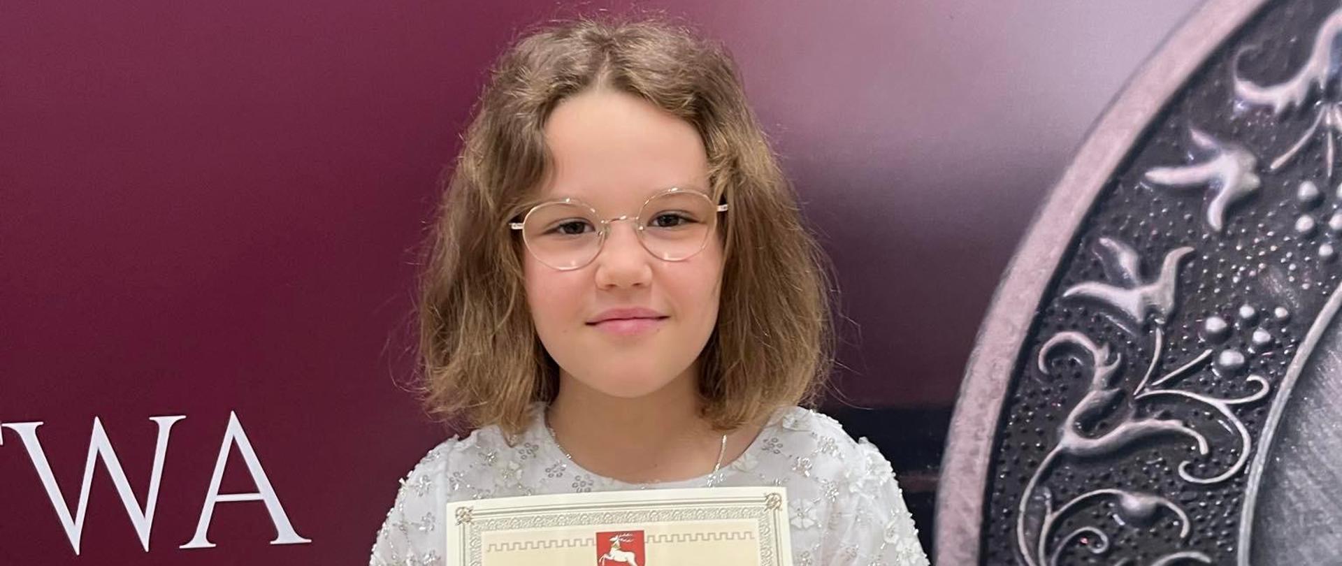 W dniu 29 kwietnia 2024 w sali widowiskowej Opery Lubelskiej odbyło się uroczyste wręczenie stypendiów dla osób zajmujących się twórczością artystyczna, upowszechnianiem kultury i opieką nad zabytkami przez Marszałka Województwa Lubelskiego. Uczennica naszej szkoły Amelia Lewkowicz z klasy IV cyklu 6-letniego gitary pani Iwony Grabowskiej otrzymała stypendium w kategorii "Najmłodszy twórca kultury". Na zdjęciu stoi z dyplomem.
