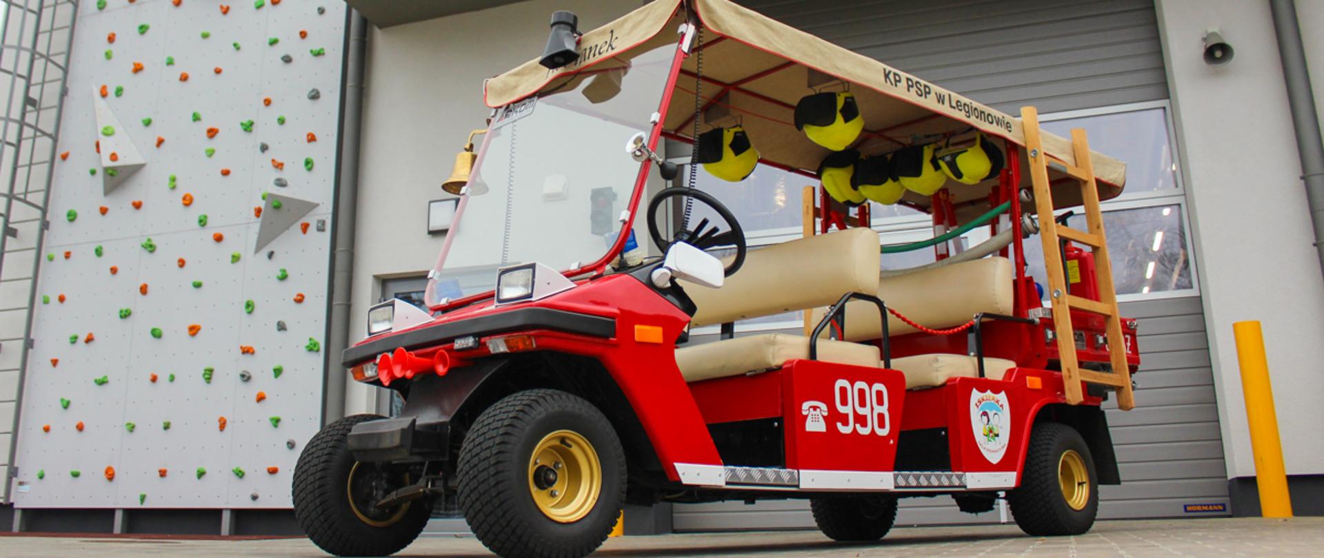 Pojazd Florianek. Zdjęcie na tle budynku KP PSP w Legionowie Pojazd czerwony melex małe kółka kremowe fotele i brezentowy daszek, pod daszkiem zawieszone żółte hełmy dla dzieci z tyłu na boku zawieszona drabinka drewniana. 