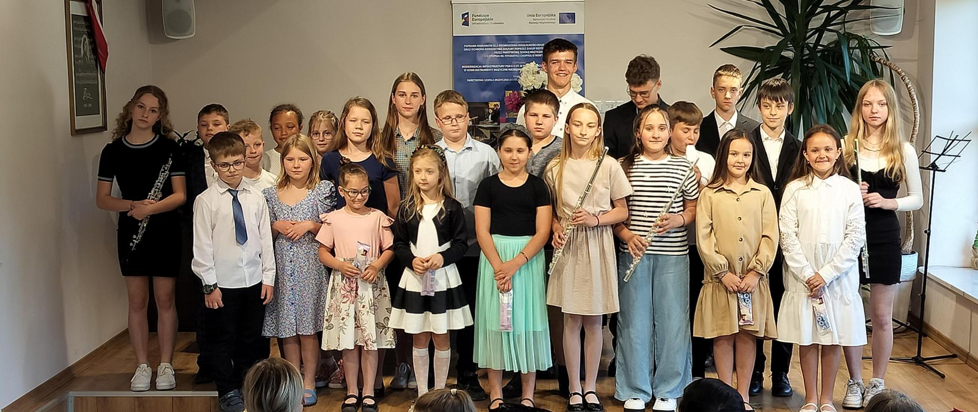 Sala kameralna, na podium stoją dzieci, uczestnicy koncertu. W rękach trzymają instrumenty oraz lody. Na ścianie po lewej stronie wisi obraz. Za dziećmi baner reklamowy zakupu instrumentów muzycznych ze środków Unii Europejskiej. Przed scena widownia.