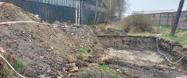 Odkopany fragment terenu. Po lewej góra piasku i w tle niebieskie blaszane budynki. Po prawej betonowy płot. Teren odgrodzony taśmą.