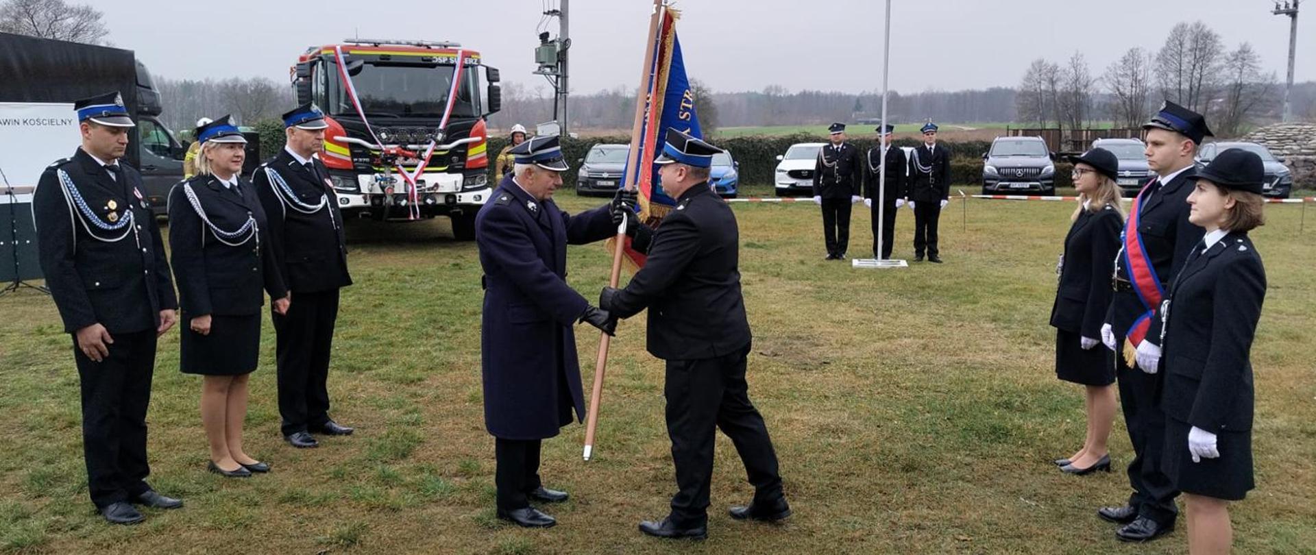 Uroczystość na trawie: dwóch strażaków w mundurach galowych przekazuje sobie sztandar. Otaczają ich strażacy OSP w mundurach, a w tle widoczny jest wóz strażacki Scania, ozdobiony biało-czerwoną wstęgą.