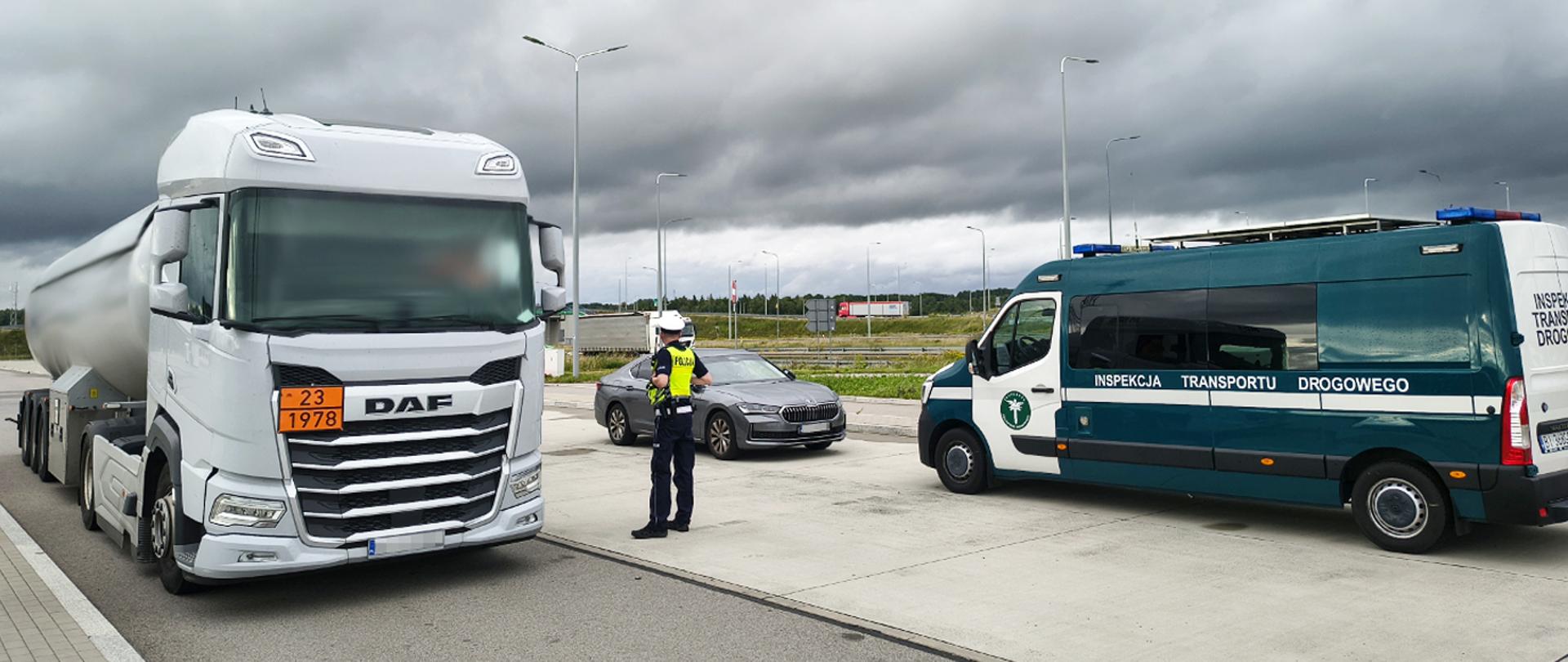 Biała cysterna z pomarańczową tablicą ADR stoi na parkingu. Obok pojazdu znajduje się funkcjonariusz oraz oznakowany samochód Inspekcji Transportu Drogowego. W tle widoczne są droga, latarnie.
