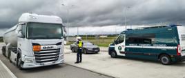 Biała cysterna z pomarańczową tablicą ADR stoi na parkingu. Obok pojazdu znajduje się funkcjonariusz oraz oznakowany samochód Inspekcji Transportu Drogowego. W tle widoczne są droga, latarnie.

