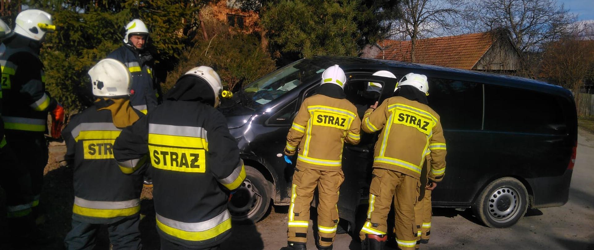 Kilku strażaków stoi obok rozbitego samochodu ciemnego koloru.