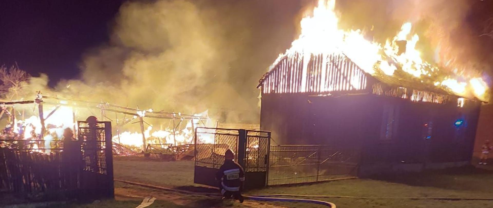 Na zdjęciu widać dwa budynki w ogniu. Jeden spłonął już niemal cały. Drugi ma w ogniu cały dach. Widać dużo dymu unoszącego się do góry. Na zdjęciu widać również strażaka, który obok bramy wjazdowej rozkłada białe węże strażackie. Jest ciemno.