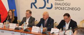 Na zdjęciu widnieje stół prezydialny za którym siedzą od lewej strony: Agnieszka Dziemianowicz-Bąk Ministra Rodziny, Pracy i Polityki Społecznej, Wiceprzewodnicząca Rady Dialogu Społecznego, Piotr Duda – przewodniczący Rady Dialogu Społecznego, Jan Klimek - Prezes ZRP, Jacek Męcina - Konfederacja Lewiatan. W tle siedzących dwie flagi: UE i Polski oraz baner z napisem Rada Dialogu Społecznego.