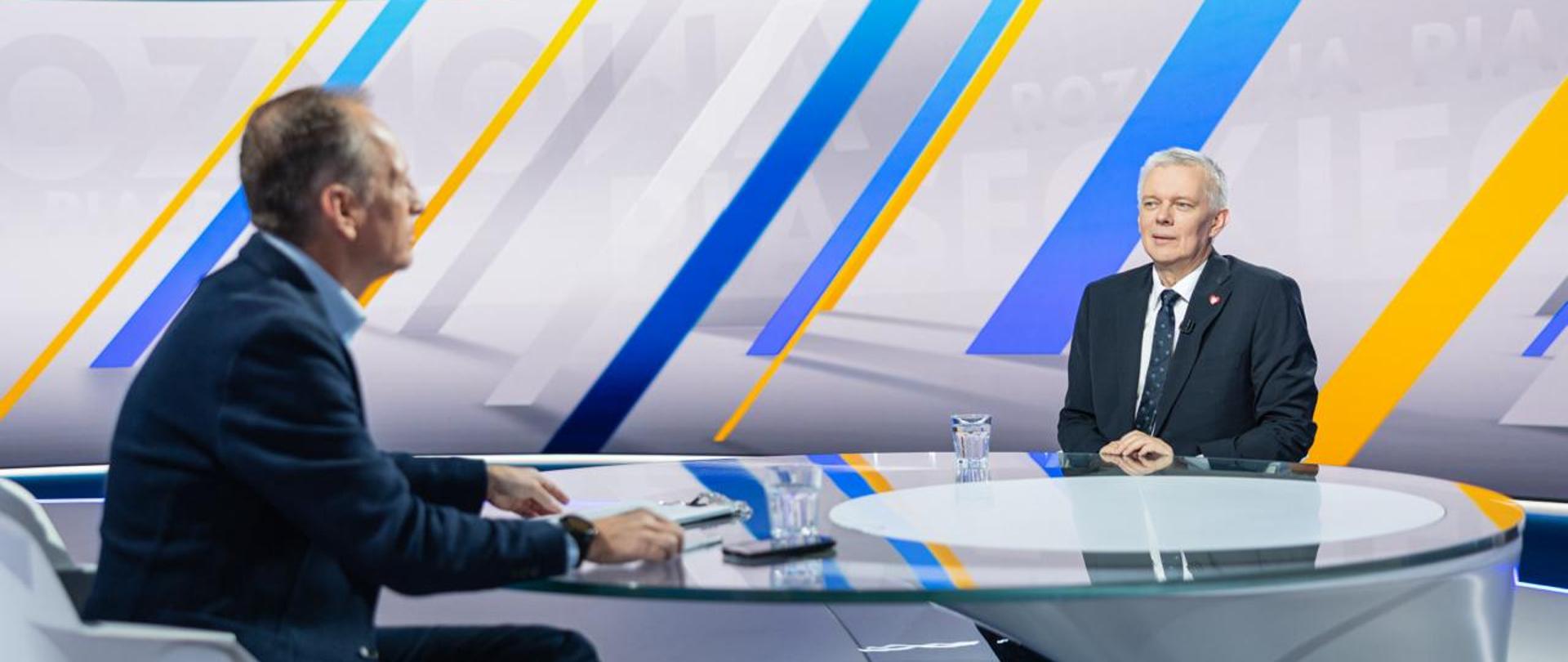 Dwie osoby przy szklanym stole. Po prawej stronie minister Tomasz Siemoniak, po lewej Konrad Piasecki. Na stole stoją dwie szklanki z wodą i telefon.