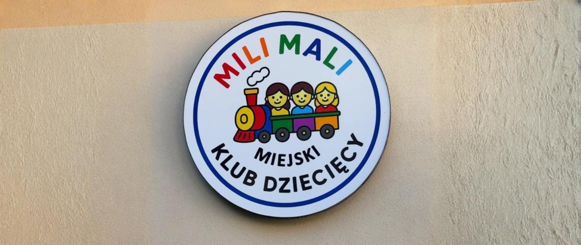 Na zdjęciu logo Miejskiego Klubu Dziecięcego „Mili Mali” - kolorowe litery MILI MALI, pod nim ciuchcia z trójką dzieci (obrazek), poniżej napis MIEJKI KLUB DZIECIĘCY.