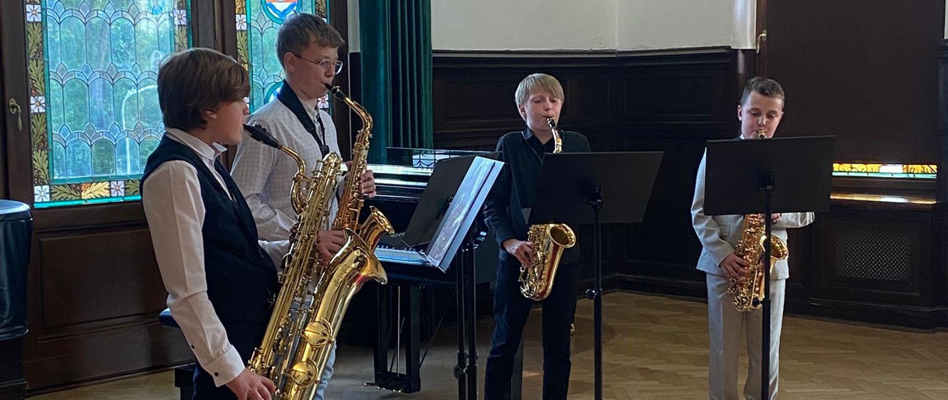 Czterech młodych uczniów gra na saksofonach w sali koncertowej Państwowej Szkoły Muzycznej w Szczecinku. Stoją na podwyższeniu z czerwonym dywanem, każdy przy pulpicie z nutami. W tle widoczne są kolorowe witraże, drewniane boazerie oraz tablica z nazwą szkoły. Występ odbywa się w eleganckim, historycznym wnętrzu.
