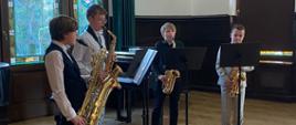 Czterech młodych uczniów gra na saksofonach w sali koncertowej Państwowej Szkoły Muzycznej w Szczecinku. Stoją na podwyższeniu z czerwonym dywanem, każdy przy pulpicie z nutami. W tle widoczne są kolorowe witraże, drewniane boazerie oraz tablica z nazwą szkoły. Występ odbywa się w eleganckim, historycznym wnętrzu.