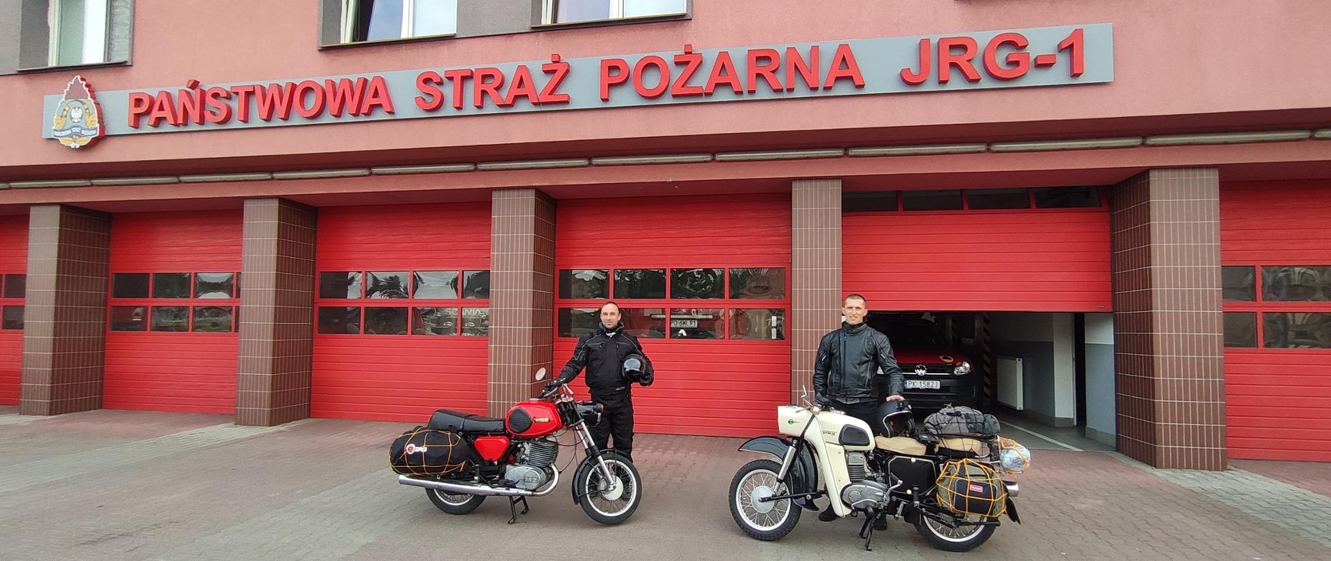 Na zdjęciu widać dwie osoby. Stoją przed budynkiem komendy PSP w Kaliszu. W tle garaże JRG 1. Zdjęcie wykonane na kostce brukowej. Przy osobach motocykle.