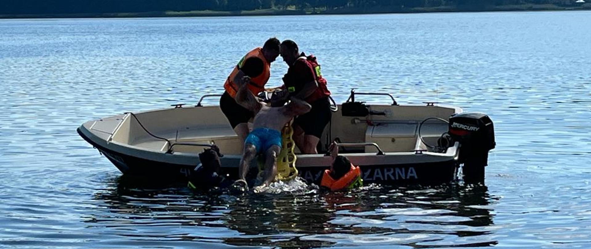 na zdjęciu czterech strażaków podejmujących osobę tonącą na łódź, w tle Jezioro Miejskie