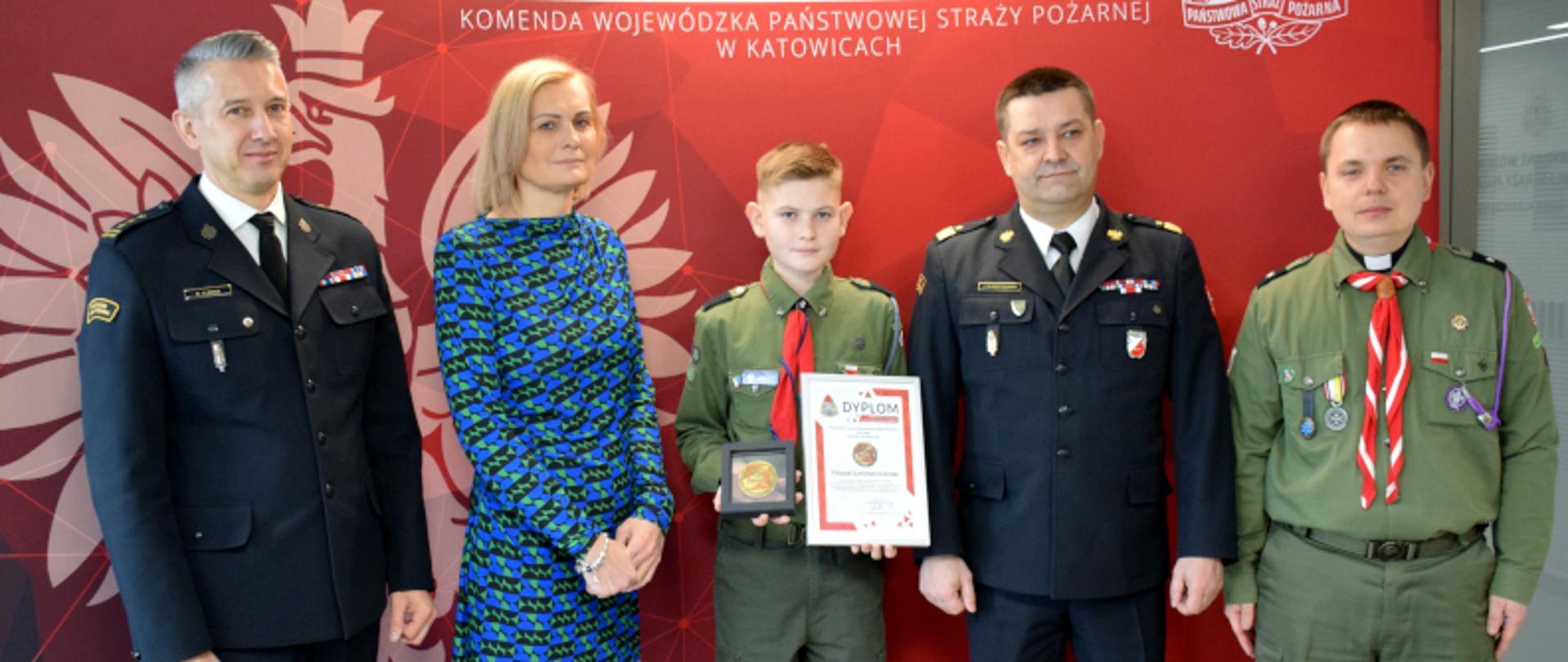 od lewej: Komendant Miejski Państwowej Straży Pożarnej w Gliwicach st. bryg. mgr inż. Roman Klecha, Filip wraz z mamą, Śląski Komendant Wojewódzki PSP nadbryg. Jacek Kleszczewski oraz duszpasterz harcerzy
