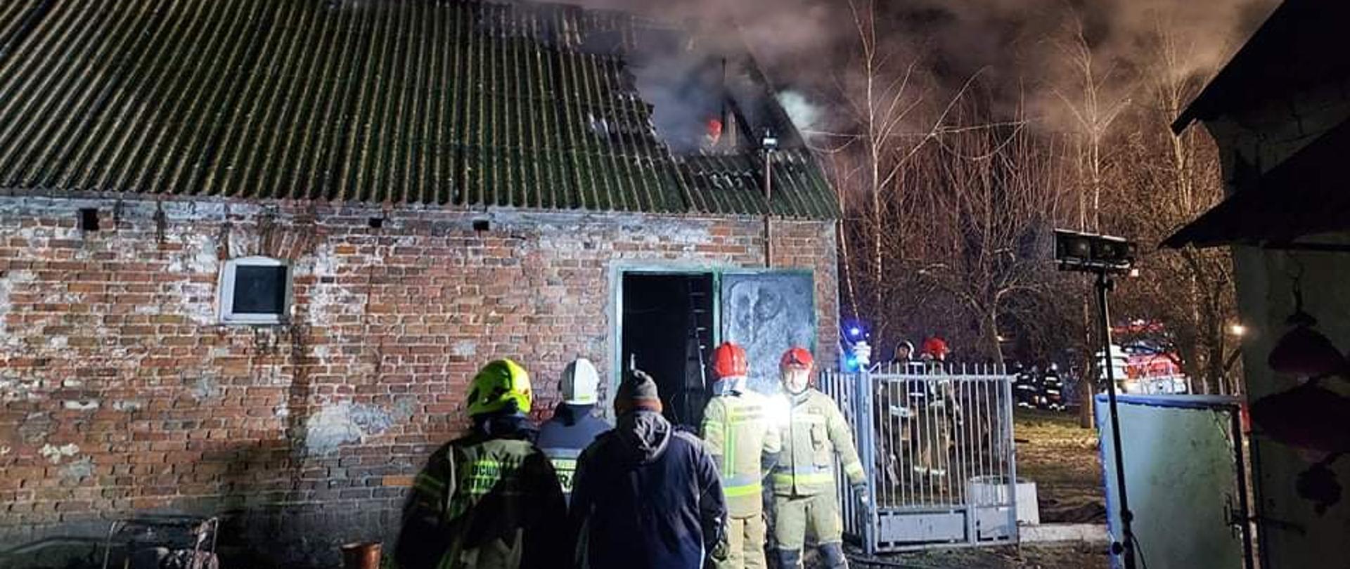 W tle budynek gospodarczy z czerwonej cegły. Częściowo nadpalona prawa strona dachu. Przed wejściem stoją strażacy