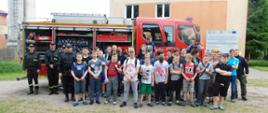 Kolejne grupowe zdjęcie zadowolonych uczestników pokazów strażackich. Na zdjęciu widoczny jest samochód straży Pożarnej wraz z uczestnikami i strażakami.