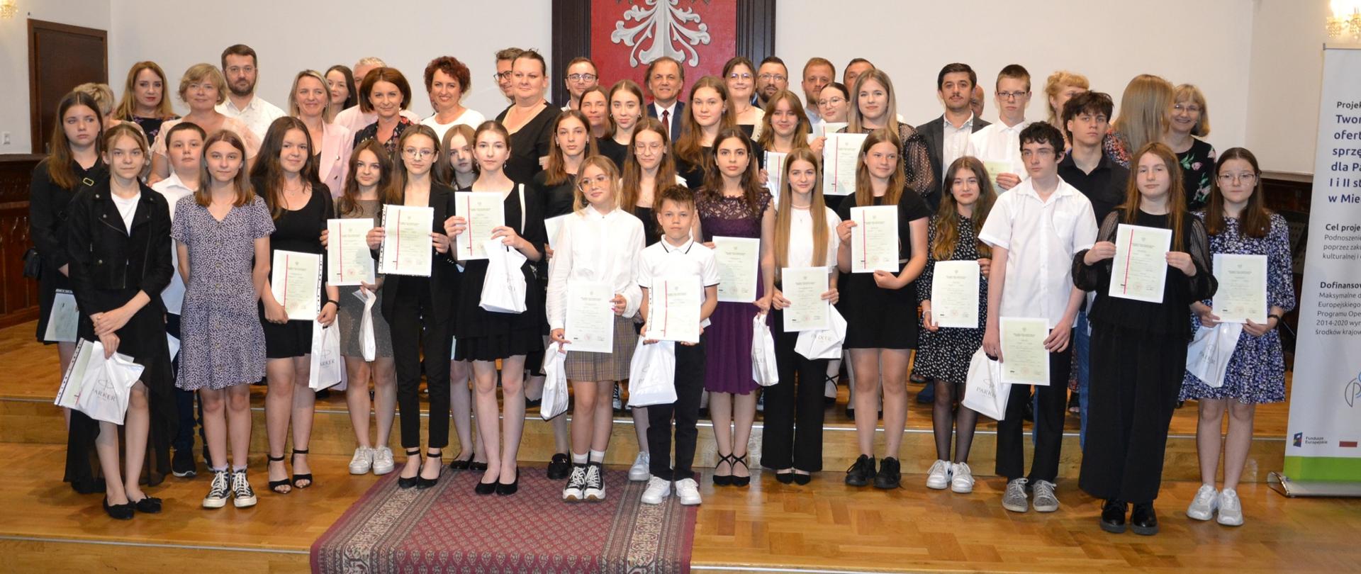 grupa uczniów i nauczycieli zgromadzona na scenie Sali Królewskiej w PSM Mielec podczas zakończenia roku szkolnego