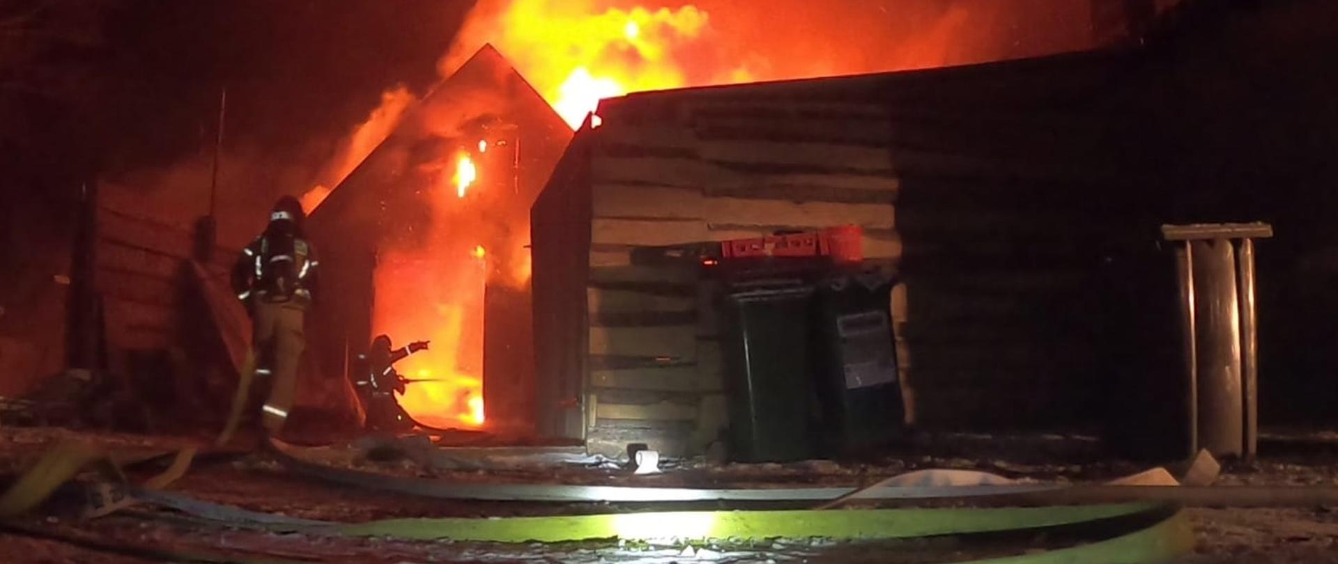 Palący się cały budynek gospodarczy. Gaszą go dwaj strażacy ubrani w ubranie bojowe z aparatami ochrony dróg oddechowych na plecach. 