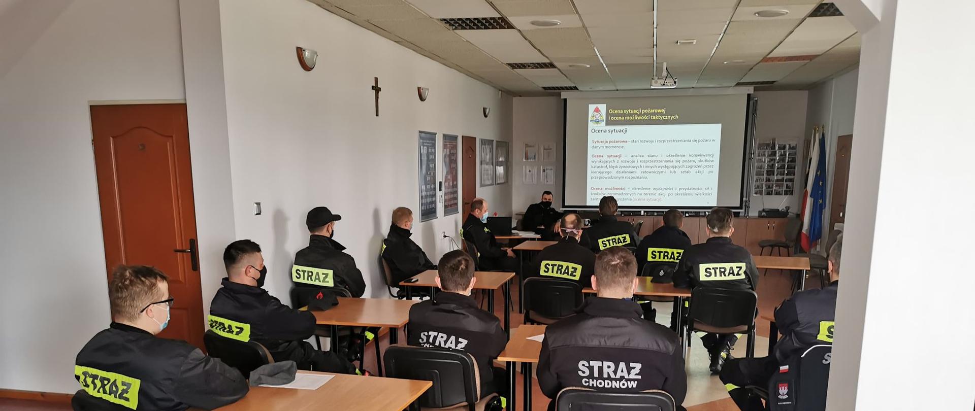 Na zdjęciu widać siedzących w sali ok. 16 mężczyzn. Wszyscy siedzą w ławkach plecami do fotografa, ale przodem do rzutnika, który znajduję się przed nimi. Wszyscy mężczyźni ubrani są w czarne strażackie stroje koszarowe z napisem STRAŻ na plecach.