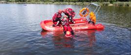 Strażacy w skafandrach wciągają na łódkę ratowniczą innego strażaka - pozoranta. W tle zarośla. 
