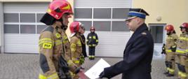 Strażacy stoją na uroczystej zbiórce. Komendant wręcza im awanse. 2 strażaków ubranych jest w ubranie specjale a 3 w mundur. 
