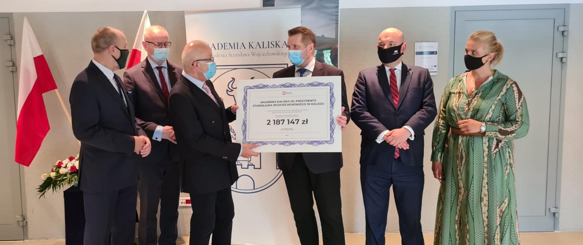 Minister Czarnek stoi w otoczeniu członków kolegium Akademii Kaliskiej, trzymając symboliczny czek - dużą kartkę z napisem 2 187 147 zł.