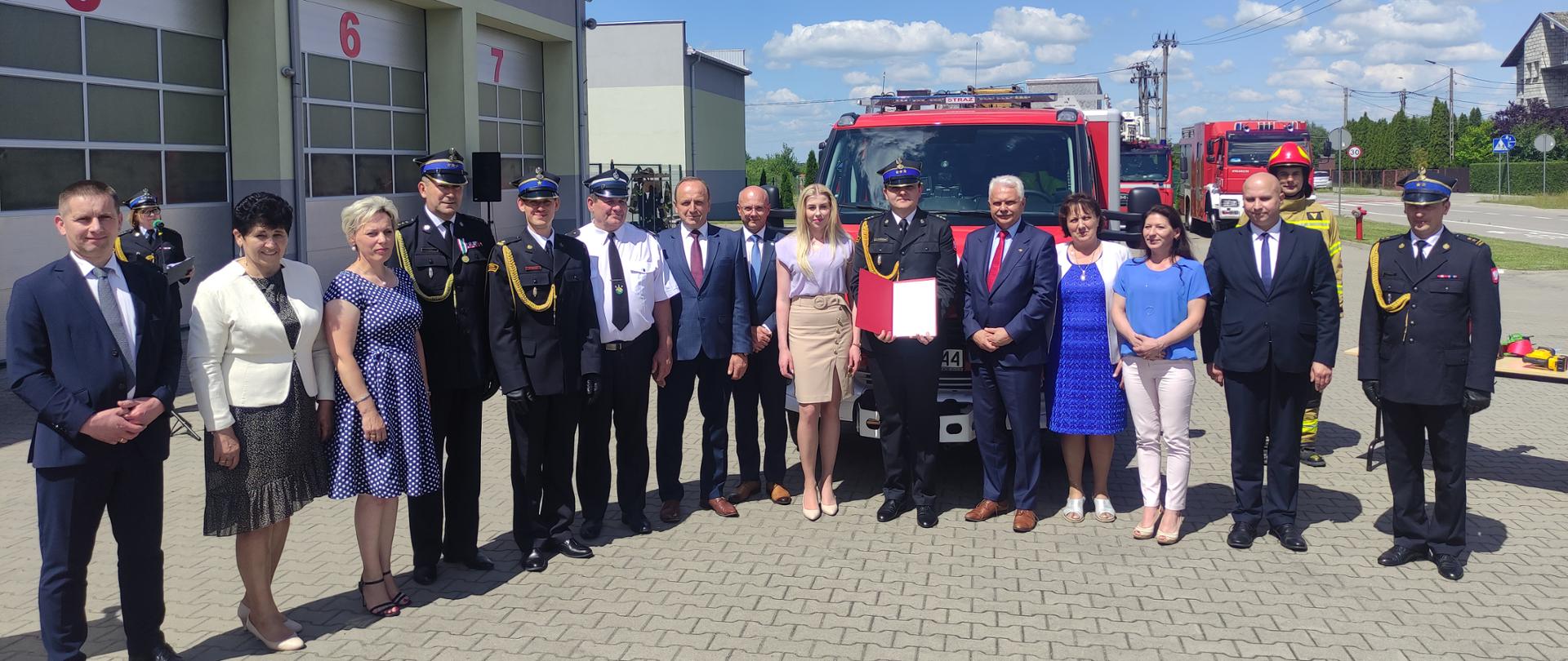 Powiatowe Obchody Dnia Strażaka 2022 r. - na placu przed strażnicą PSP w Sokołowie Podlaskim , trwają uroczystości. Goście stoją na zbiórce w szeregu przed czerwonym samochodem strażackim.