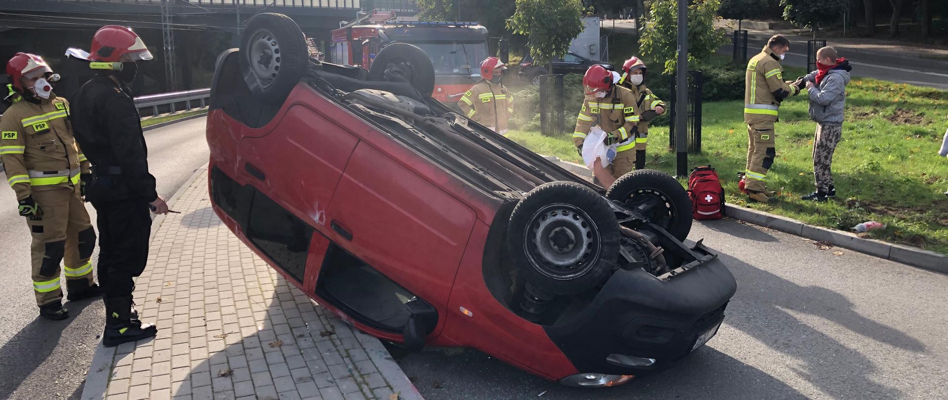 Zdjęcie przedstawia przewrócony na dach samochód fiat panda. Obok działania powadzą strażacy, rozsypują sorbent oraz udzielają pomocy przedmedycznej poszkodowanemu. Na zdjęciu widać również samochód rtowniczo-gaśniczy JRG Sopot 