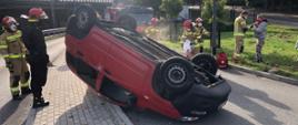 Zdjęcie przedstawia przewrócony na dach samochód fiat panda. Obok działania powadzą strażacy, rozsypują sorbent oraz udzielają pomocy przedmedycznej poszkodowanemu. Na zdjęciu widać również samochód rtowniczo-gaśniczy JRG Sopot 