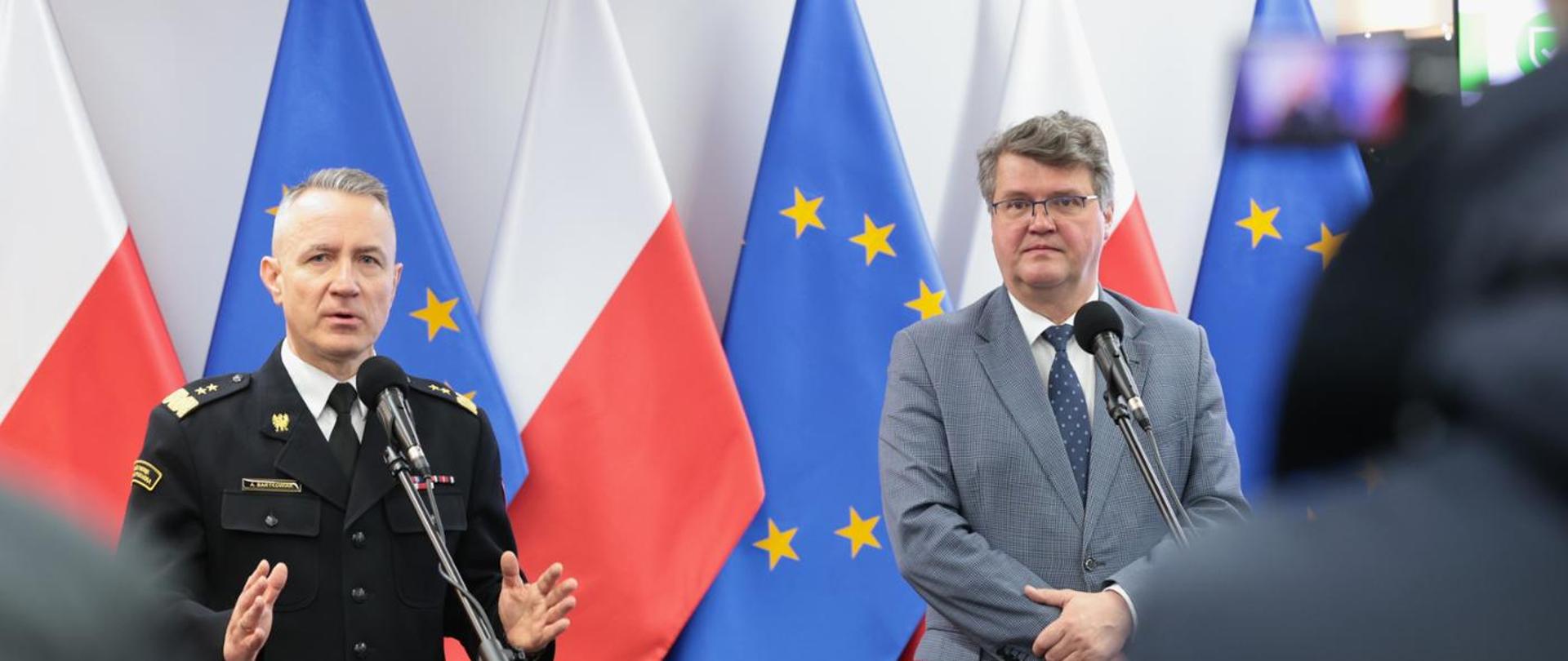 Na zdjęciu na tle flagi RP i UE przy mikrofonach stoi wiceminister Maciej Wąsik i komendant Główny gen.bryg. Andrzej Bartkowiak podczas konferencji prasowej