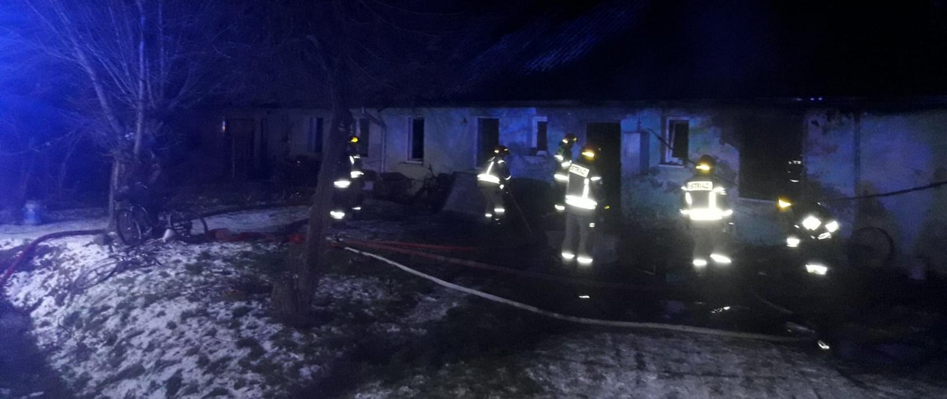 Zdjęcie wykonane wieczorową porą na miejscu działań gaśniczych. Strażacy w założonych ubraniach specjalnych z odblaskowymi taśmami które odbijają światło oraz czerwonych hełmach. Na ziemi leżą linie wężowe z wodą.