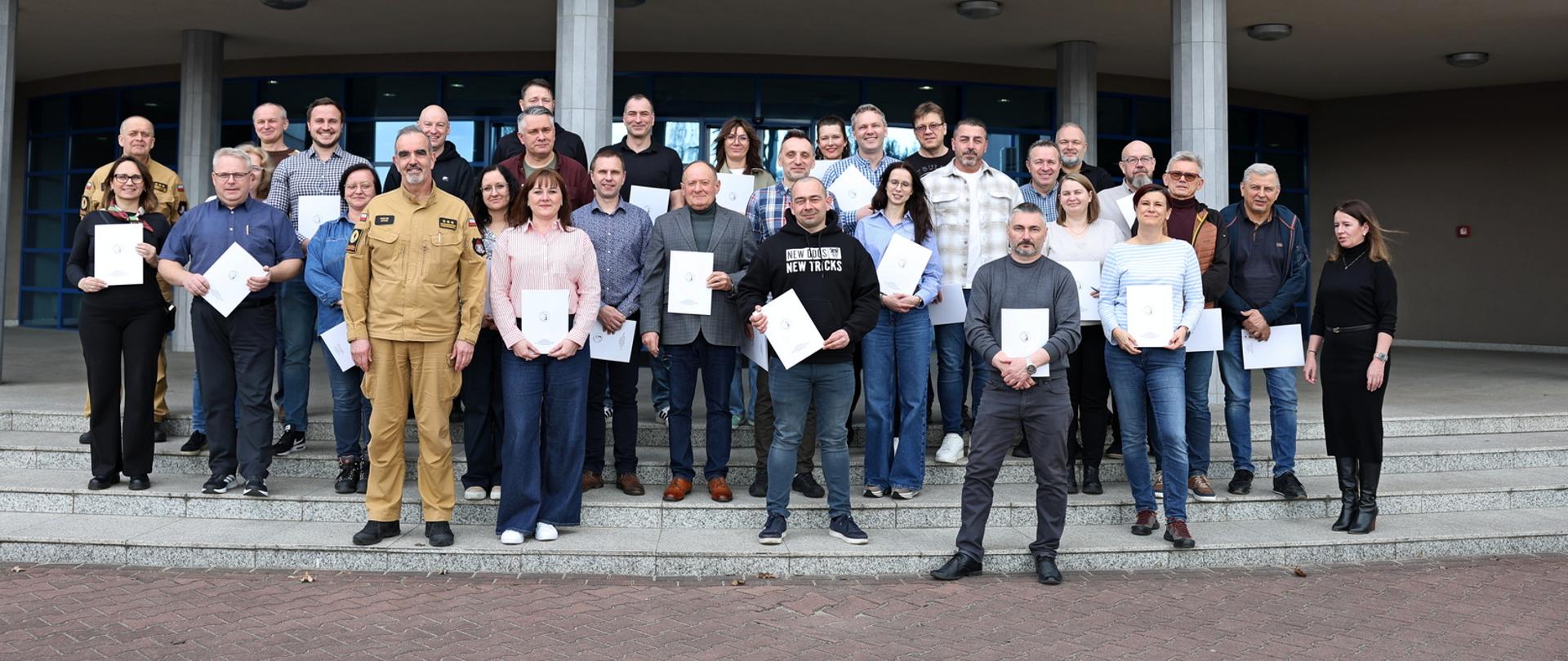 Pamiątkowe zdjęcie grupowe uczestników szkolenia dla inspektorów ochrony przeciwpożarowej (30 osób) w towarzystwie Zastępcy Komendanta Centralnej Szkoły PSP, oficera CS PSP oraz kobiety. Uczestnicy trzymają w dłoniach dokumenty potwierdzające ukończenie szkolenia.
