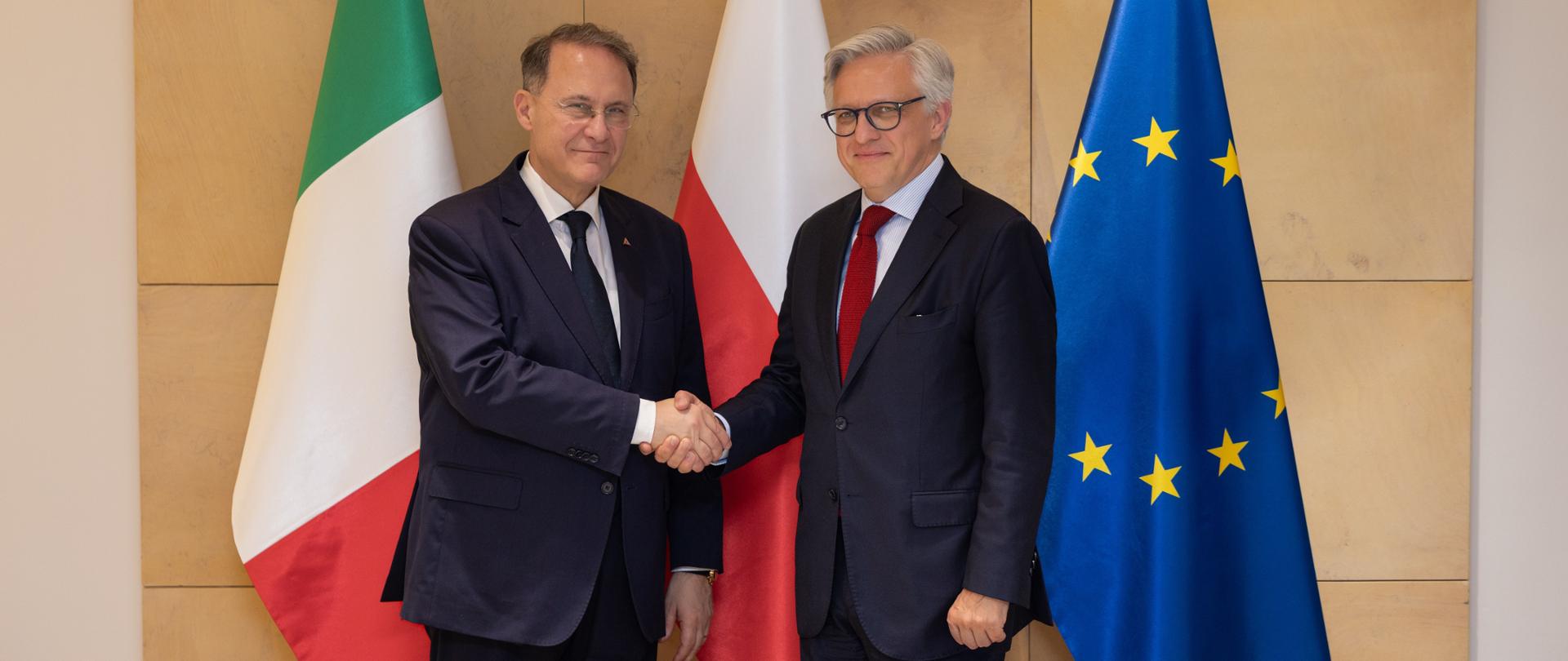 Two men in dark suits shake hands in front of the flags of Italy, Poland, and the European Union - Deputy Minister of Foreign Affairs and International Cooperation of the Italian Republic Edmondo Cirielli and Undersecretary of State Artur Harazim. The meeting takes place against a wall displaying the Polish coat of arms, highlighting the official diplomatic nature of the event.